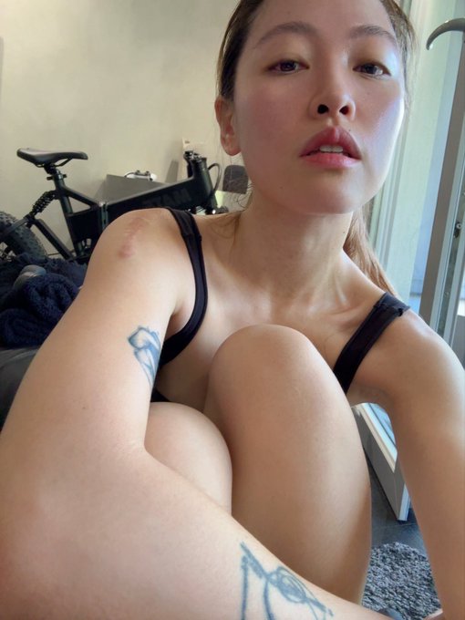 A selfie taken by a young woman with blonde hair wearing a black sports bra, revealing a tattoo on her forearm, with a stationary bike visible in the background.
