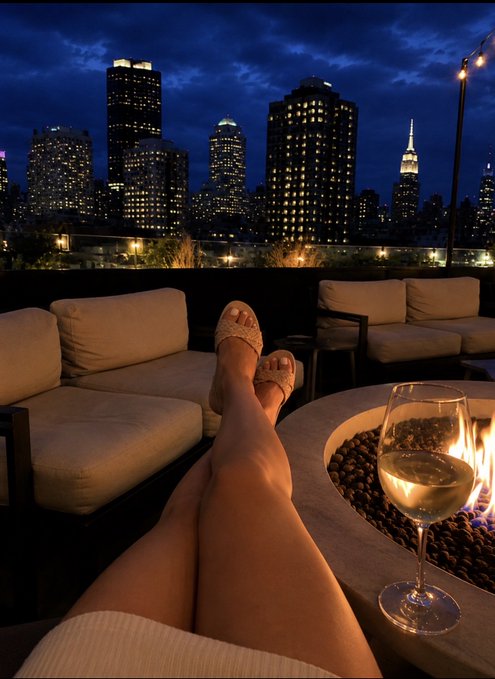 The image shows a woman sitting on a couch with her legs crossed while wearing white sandals. She has her feet resting on the arm of the couch, and there are two wine glasses placed next to her. The scene takes place at night, with a city skyline visible in the background. The post seems to be about the excitement or hype surrounding a particular event or situation, as it says "HYPE up."