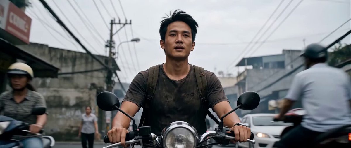 The image shows a man riding a motorcycle down a street with buildings in the background. There are other people walking nearby, indicating that this scene takes place in Ho Chi Minh City, Vietnam. The photo captures the essence of city life, showcasing the fast-paced nature of urban living and the various modes of transportation available to navigate through it.