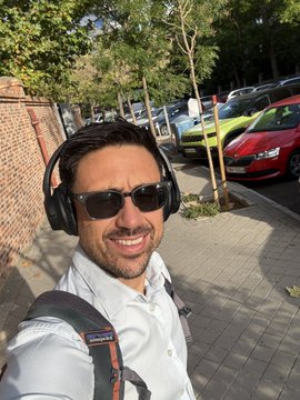 A smiling man wearing sunglasses, black headphones, and a light blue shirt takes a selfie on a sunny street with parked cars in the background.