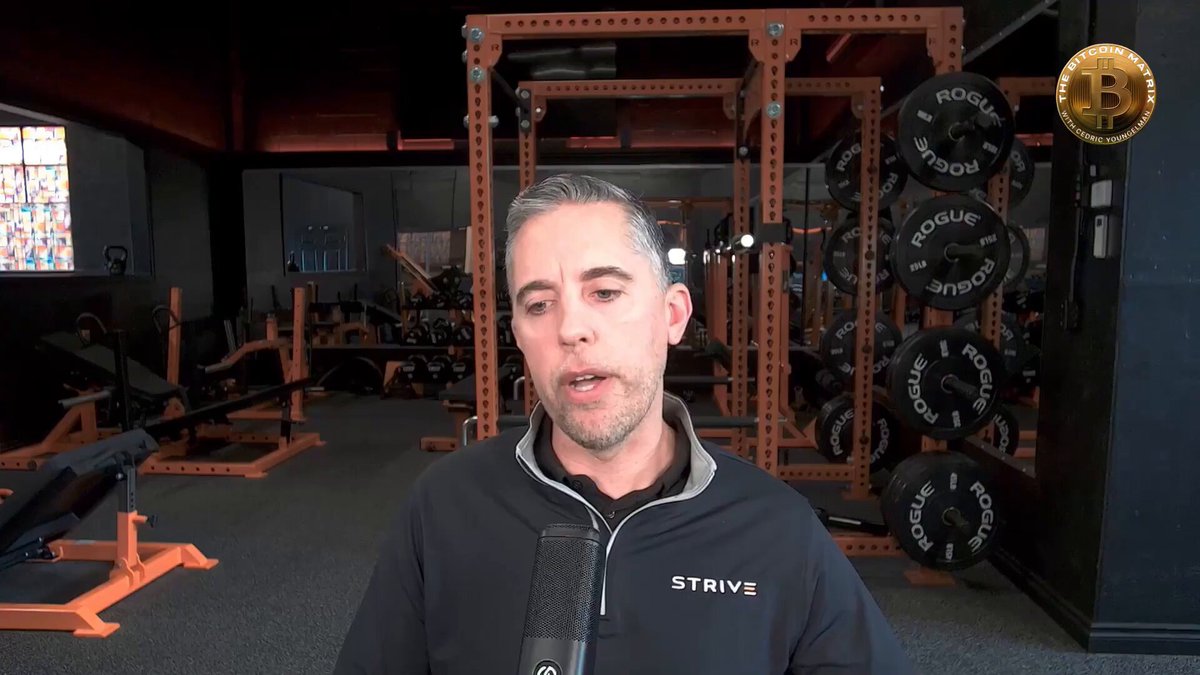 The image shows a man wearing a black jacket, standing in front of a weight rack at a gym. He appears to be talking into a microphone or recording device while surrounded by various exercise equipment such as dumbbells and barbells. The background features an orange wall with a window that allows natural light to enter the room.

The image also contains text that reads "Just In: Strive $ASST CEO Matt Cole called Saylor and Phong Le " which refers to the man's name, Matt Cole, as the CEO of Strive Fitness. The post also mentions that Strategy $MSTR will be the largest company in the world by the time they retire from their current positions at age 65.