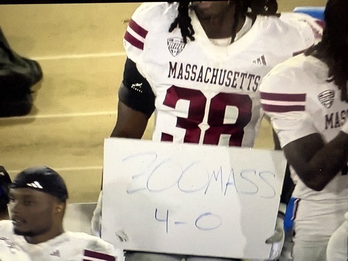 A UMass football player wearing jersey number 38 sits on a bench holding a handwritten sign that reads 'BC MASS 4-0'.