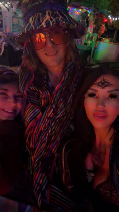 Three people pose together at what appears to be a colorful festival or bar setting, with the central figure wearing a fuzzy hat and orange-tinted sunglasses, while the woman on the right has a decorative forehead piece.