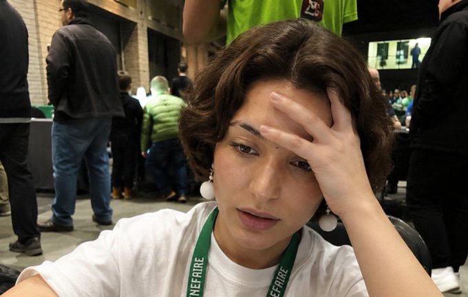 A young woman with wavy brown hair and white pom-pom earrings sits looking exhausted with her hand on her forehead, wearing a green lanyard that reads "BENEFARE." The image humorously captures the weariness of staying up late, matching the caption's complaint about waiting for Moonpay.