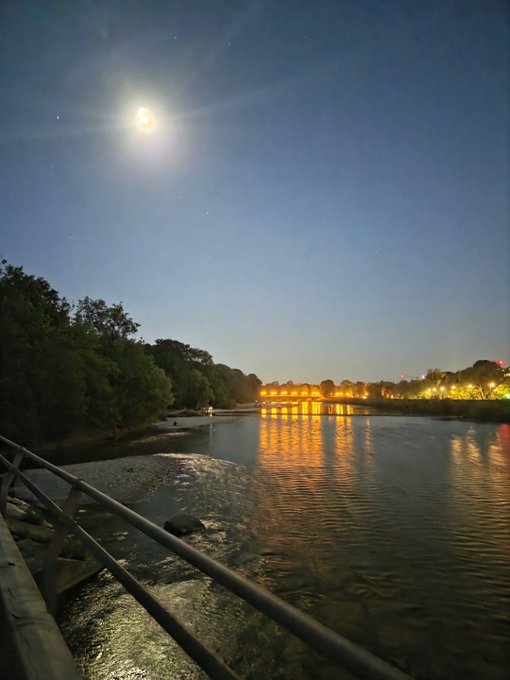 A nighttime scene over a river with the moon shining brightly above, illuminating a lit bridge in the distance and reflecting on the water below.