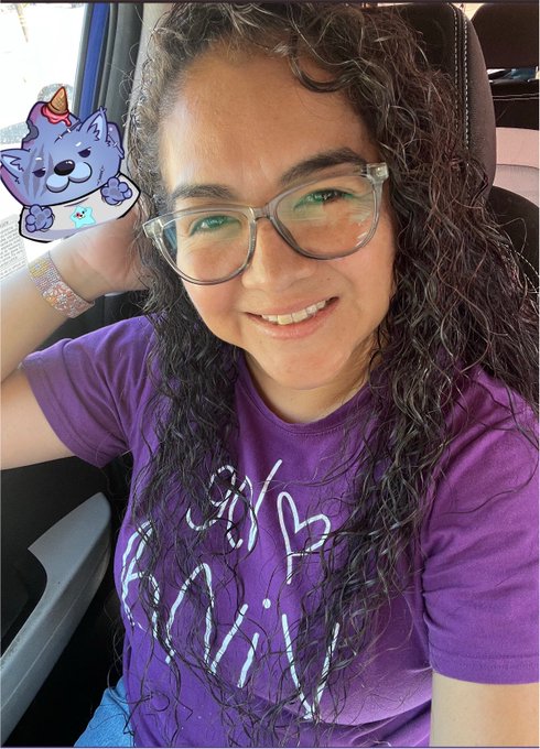 A selfie of a woman with curly hair and glasses wearing a purple t-shirt, sitting in a car with a cartoon cat sticker overlay on the image.