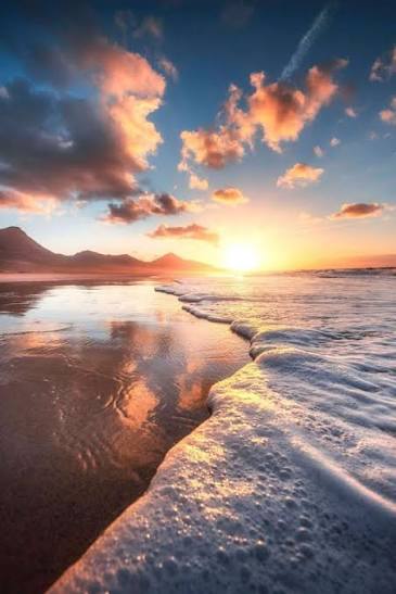 The image shows a beautiful beach scene at sunset, with the sky filled with clouds and the ocean waves crashing onto the shore. There are mountains visible in the background, adding to the picturesque view. The photo has been captioned "Another week, another reminder that real utility matters. A lot of Web3 apps focus on speculation, but @timesoulcom is taking a different approach by connecting blockchain incentives with everyday wellness habits." This message emphasizes the importance of incorporating healthy routines into daily life and highlights how the app encourages users to do so through its unique approach.