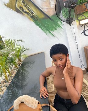 A casual portrait of a young woman with short hair sitting on a chair outdoors, wearing a brown top and looking at the camera with her hand near her face.