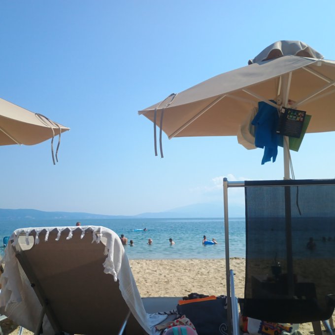 A sunny beach scene with beige sun umbrellas, a beach chair draped with a white towel, and people swimming in calm blue water with mountains visible on the horizon.