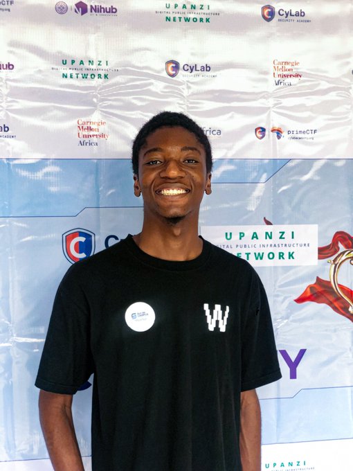 A portrait photo of a smiling young man wearing a black 'W' (West Virginia University) t-shirt, standing in front of a sponsor backdrop featuring logos for Upanzi Network, CyLab, Nihub, and Carnegie Mellon University Africa.