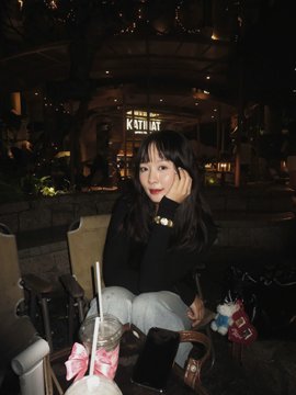 A young woman with long dark hair and a bow tie sits at a nighttime outdoor café table under a sign reading "K-STYLE," with drinks and bags placed in front of her. The cozy, stylish setting reflects the post's weekend leisure vibe as she invites followers to check in and support her new episode of the Vietnamese crypto news series.