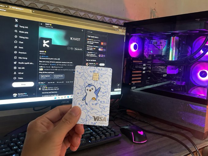 The image shows a person holding up a Visa card that has a penguin design on it. The card is being held above a computer monitor, which displays a website with a blue background featuring a penguin icon. The text accompanying the image reads "Some cards are just payment methods. Some cards tell the story of an entire journey. From countless hours in front of the screen following markets, chasing opportunities, engaging with the community, and growing alongside KAST to finally holding the Pudgy Card in my hand." This suggests that the person is using their Visa card for online purchases or transactions related to penguin-themed items or experiences.