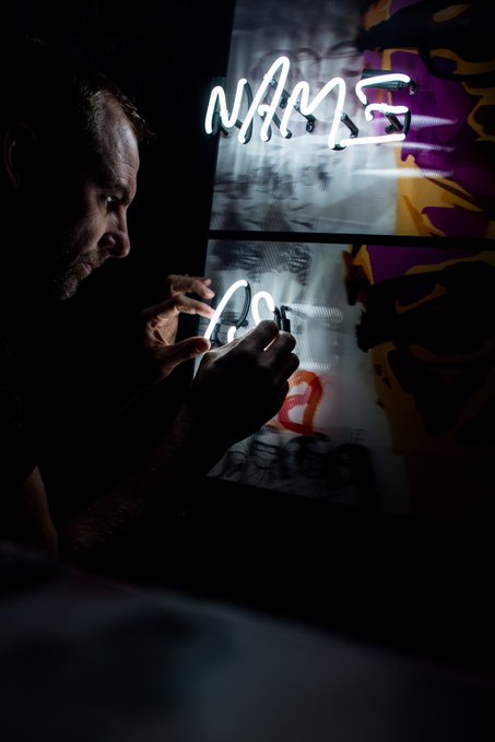 A bearded man in a dark setting carefully working on illuminated neon signage that reads 'NAME' and 'DG' over colorful printed backdrops.