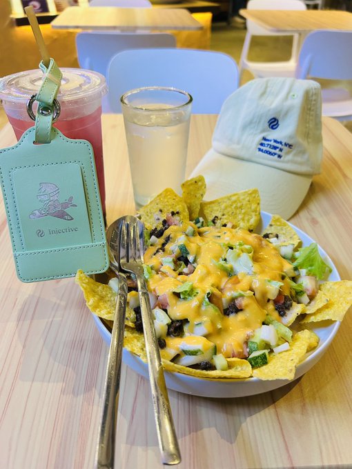 The image shows a table with a bowl filled with food, including nachos topped with cheese and other toppings. There are also two cups - one containing a drink and another empty. A green tag or lanyard can be seen on the table as well. The caption reads "FLASH TASK ALERT FOR WEEK 2 $INJ SQUAD CHALLENGE i feel hungry so i just grab a bite".