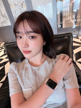 A young woman with short brown hair wearing a light-colored button-up top and a white smartwatch, posing indoors in front of a window with a patterned floor visible.