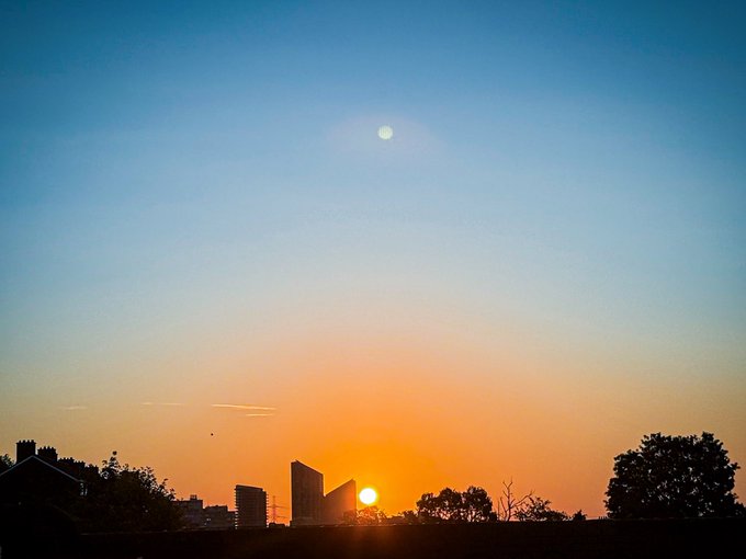 A serene sunrise cityscape with the sun peeking over silhouetted buildings beneath a blue sky featuring a visible moon. The photo captures the photographer's artistic eye, paired with their reflective message apologizing for their absence while reaffirming their dedication to photography and the NFT community.