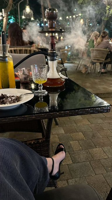 A person's legs crossed under a dark wicker table at an outdoor lounge at night, with a hookah emitting smoke, drinks including orange juice, and other patrons visible in the background.