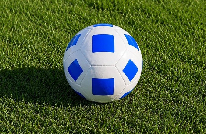The image features a soccer ball with blue and white squares, placed on a green grass field. The background appears to be a plain or empty area, which contrasts with the vibrant colors of the soccer ball. The focus of the image seems to be on the soccer ball itself rather than any additional elements in the scene.