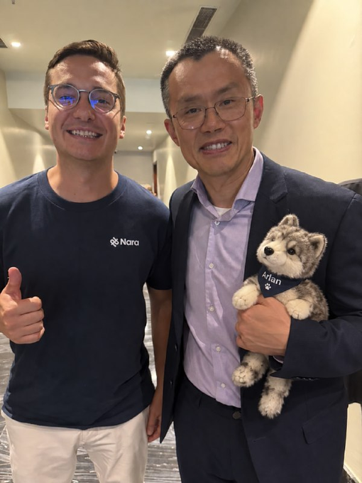 Two smiling men pose for a photo indoors; the one on the left wears a navy 'Nara' t-shirt and gives a thumbs up, while the one on the right wears a suit jacket and holds a stuffed husky dog wearing a shirt labeled 'Aidan'.