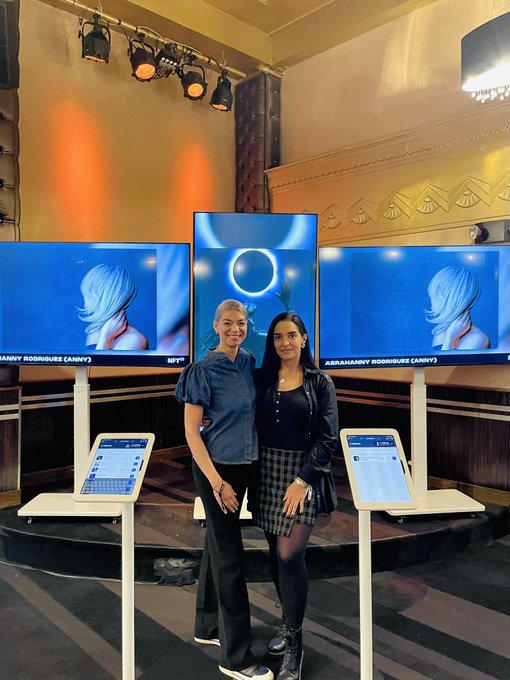 Two women pose together in front of three digital display screens labeled 'ABIRAHAMNY RODRIGUES (ANNY)' showing virtual avatars, with two tablet kiosks on stands in the foreground.