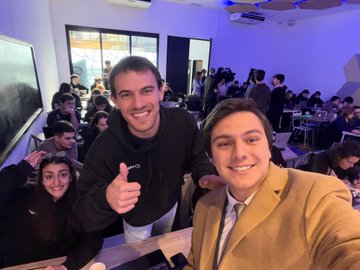 Two smiling young men pose for a selfie in a crowded room, one in a dark sweater holding a small cup and making a peace sign, while the other wears a tan blazer.