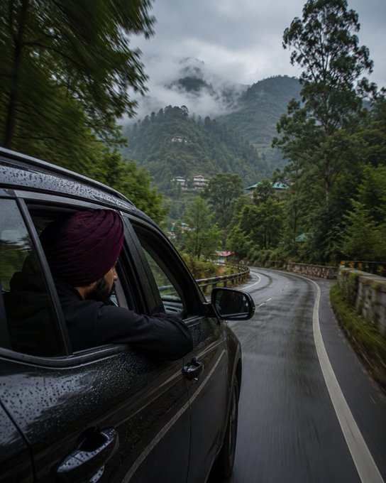 A man wearing a maroon turban looks out the window of a moving car traveling along a winding mountain road surrounded by misty green hills.