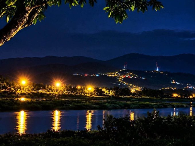 A nighttime scenic view of a riverside town with illuminated street lights reflecting on the water and mountain silhouettes in the background.