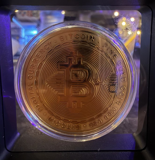 A close-up of a physical Bitcoin commemorative coin in a copper/bronze finish, encased in protective plastic, with text reading 'DIGITAL CURRENCY · BITCOIN · FINE COPPER' and '999 FINE COPPER'.