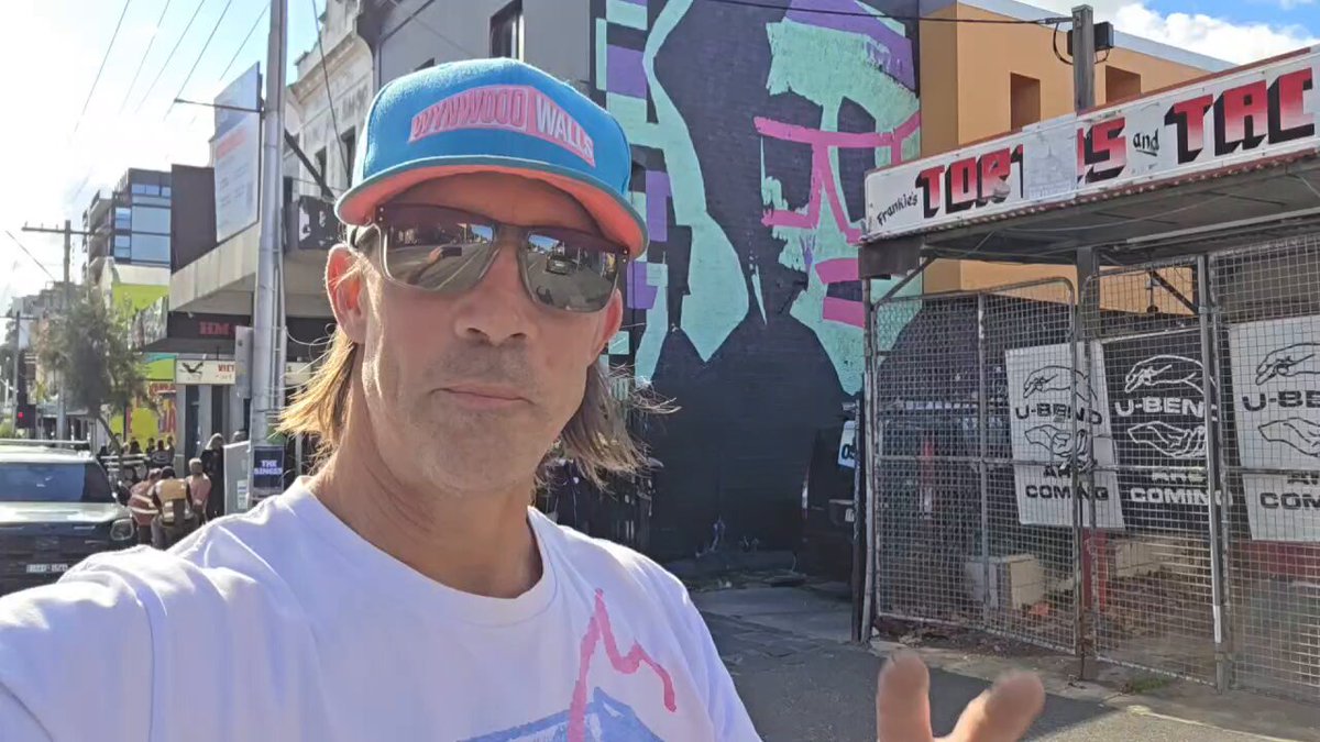 The image features a man wearing sunglasses and a baseball cap standing next to a building with graffiti art on its side. The man appears to be posing for a photo, possibly at a street fair or event. In the background, there are several cars parked nearby, indicating that this location is likely in an urban setting. The post includes text about the true crypto gallery and mentions "GT" and "Bigger and Better things family." This suggests that the man's photo might be related to a street fair or similar gathering where various businesses and attractions come together.