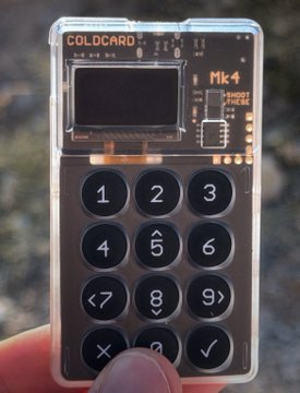 A hand holds a Coldcard Mk4 Bitcoin hardware wallet, with the keypad buttons including numbers 1-9, directional arrows, and symbols like "<7", "9>", "X", and "✓" clearly visible. The image illustrates the post's reference to the device, though it should be noted that no verified hack compromising Coldcard wallets has been publicly documented.
