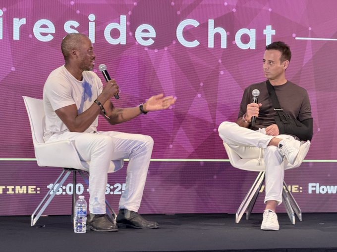 Two men sit in white chairs on a stage holding microphones during a 'Fireside Chat' event, with a time of '16:00 - 16:25' visible in the background.