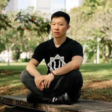 A young man wearing glasses and a black t-shirt sits cross-legged on a wooden platform in a park, with trees and greenery in the background.