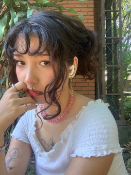 A young woman with dark wavy hair poses near a brick wall, wearing a white ruffled crop top, a pink beaded necklace, and rings, with tattoos visible on her arm and hand resting near her face.