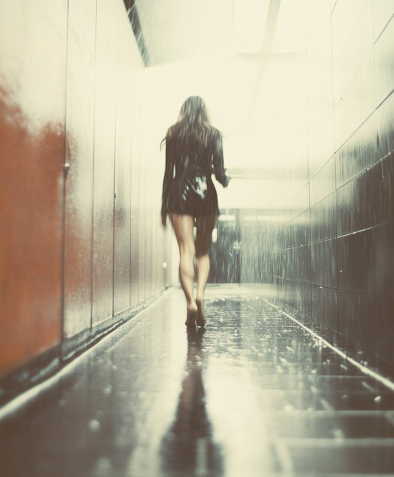 A woman in a black dress walks barefoot down a wet, dimly lit corridor as bright light pours in from the end, with rain visible on the right side.