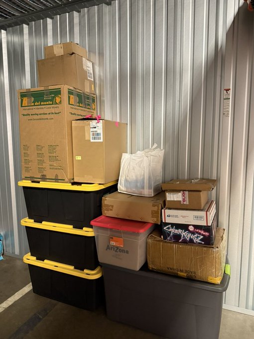 A pile of boxes and storage bins stacked in what appears to be a storage unit, including Amazon packages, an "ArtZone" container, and a box marked "yourdm" alongside NFT-branded merchandise. The caption playfully mocks the dwindling value and storage burden of 2021-era NFT merch, suggesting these boxes of digital collectibles have been relegated to a self-storage unit.