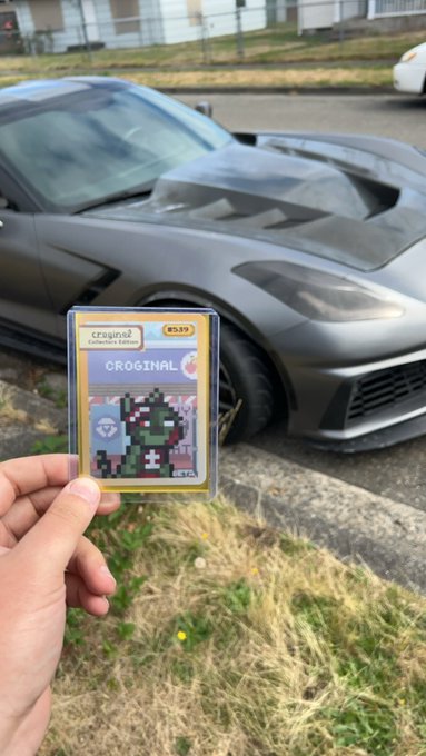 A hand holding up a Pokémon trading card labeled 'CROGINAL' (#39) in front of a matte gray Chevrolet Corvette sports car parked on the street.