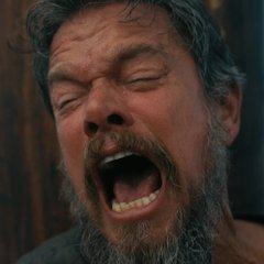 A close-up of a bearded man with graying hair laughing or shouting with his eyes closed and mouth wide open.