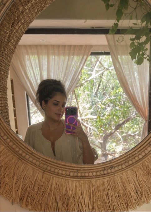 A woman takes a mirror selfie with her phone in a room with sheer curtains and a view of trees, wishing everyone a "Good morning." The bohemian-style round mirror with a fringed frame reflects her casual, natural morning moment.