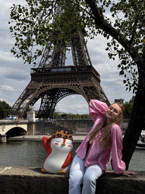 A young woman in a pink sweater poses next to a Moogle plushie (from Final Fantasy) with the Eiffel Tower in the background.