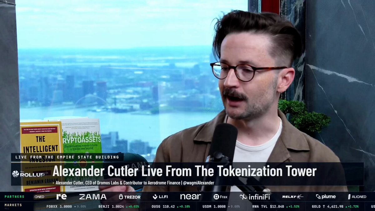 The image features a man wearing glasses, talking into a microphone while standing next to a window overlooking a cityscape. The background of the image includes text that reads "Alexander Cutler Live From The Bitcoin Bubble" and "The Bitcoin Bubble". This suggests that the man is discussing or explaining something related to the Bitcoin bubble, possibly its rise and fall in popularity or value over time.