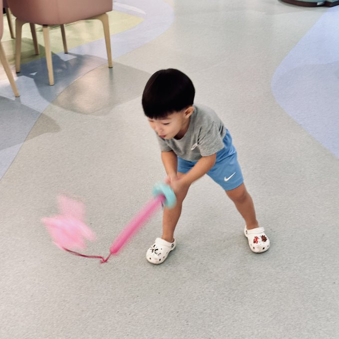 A young boy in a gray t-shirt, blue Nike shorts, and white Crocs playfully swings a pink fuzzy toy on the floor of a room.