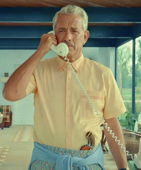 A silver-haired man in a yellow short-sleeved shirt and blue sarong stands indoors, holding a white rotary phone to his ear with a concerned expression, while a gun is tucked into his waistband.