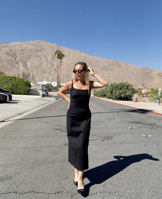 A woman in a black sleeveless dress and sunglasses standing in the middle of a desert road with mountains and palm trees in the background, casting a long shadow in the sunlight.