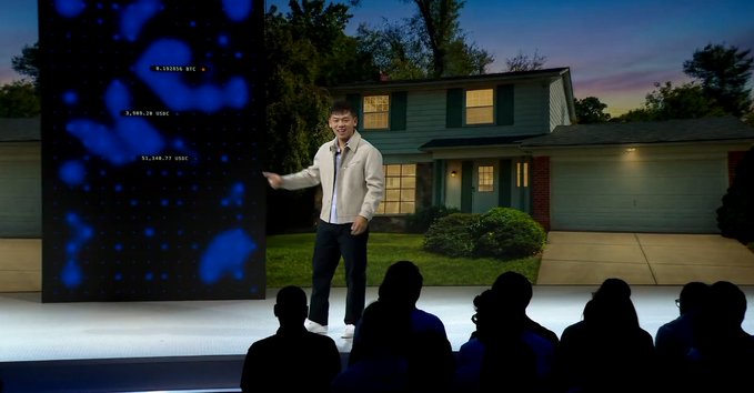 The image features a man standing in front of a house, giving a speech or presentation to an audience. The audience consists of several people who are attentively listening to him. The setting appears to be outdoors, with a grassy area visible behind the speaker. The scene is likely part of a larger event or gathering related to cryptocurrency and real estate.