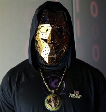 An anonymous figure wears a gold geometric mask and a black hoodie featuring a "TRUMP" logo alongside an American flag pin, evoking the mysterious identity often associated with crypto whales. This image of a concealed, wealthy-seeming figure visually captures the enigma of "Shibtoshi," the unknown on-chain trader who accumulated 103.33 trillion SHIB tokens for a paper gain of $8.9 billion without ever cashing out.