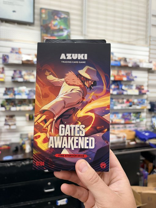 A hand holding an 'AZUKI TRADING CARD GAME GATES AWAKENED FIRE STARTER DECK' box in what appears to be a hobby or game store.