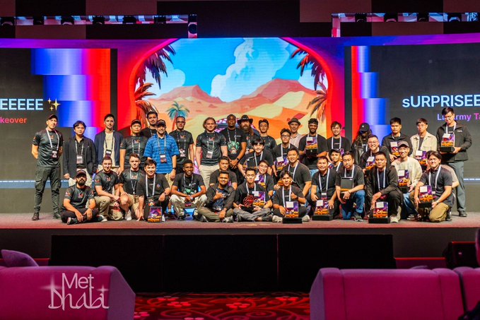 The image shows a large group photo at the Meteora hackathon event, with participants gathered on stage displaying "Met Dhālī" branding on the front of the stage and "sURPRISE" text visible on the backdrop screens behind them. Many attendees are holding awards or certificates, illustrating the speaker's joy at connecting with and positively impacting the community of builders at this event.