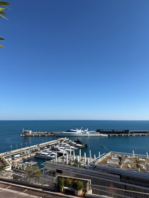An elevated view of a sunny marina featuring numerous white yachts docked alongside a pier, with a large luxury superyacht anchored near a breakwater in the blue ocean.