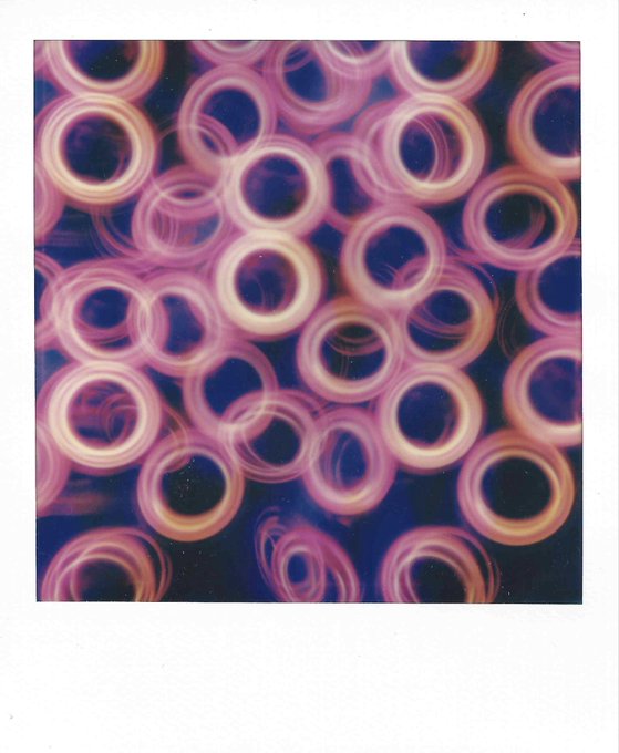 An artistic photograph with a Polaroid-style frame showing abstract, overlapping pink and white glowing circular light trails against a dark purple background.
