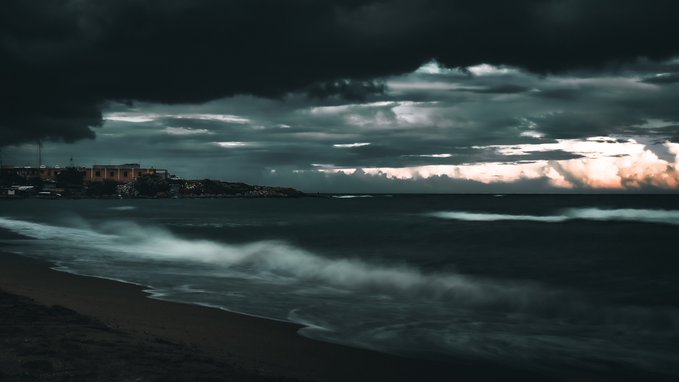 A dramatic, dark coastal landscape features a sandy shore with crashing waves under a brooding sky heavy with storm clouds, with a faint silhouette of a coastal town visible on the distant left horizon.